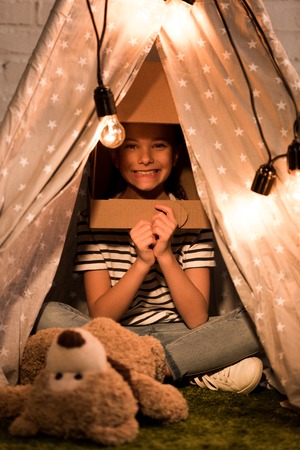 Excited kid in cardboard helmet sitting in wigwam with smileの写真素材