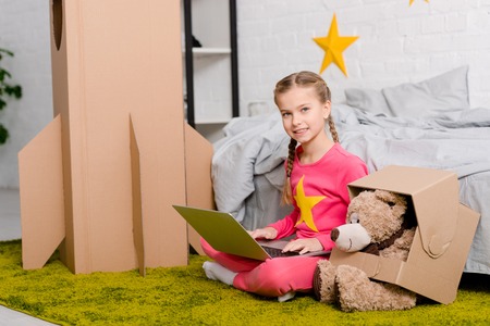Smiling kid with teddy bear sitting on carpet and using laptop in bedroom