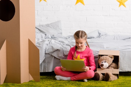 Pleased kid sitting on carpet with teddy bear and using laptop
