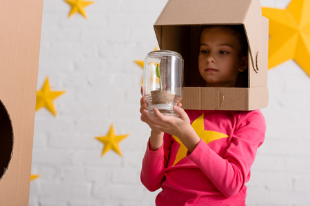 Curious child in cardboard helmet holding jar with plantの写真素材