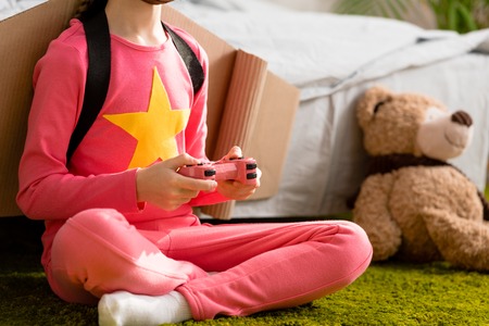 Partial view of kid in pink clothes sitting on carpet with joystick