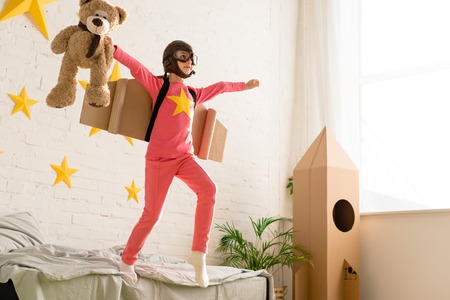 Active child with cardboard wings posing on bed with teddy bear