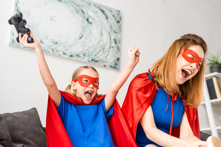 Mother and daughter in red masks and cloaks playing video game at homeの写真素材