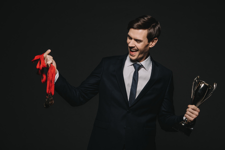 cheerful businessman looking at medals and holding trophy isolated on blackの写真素材