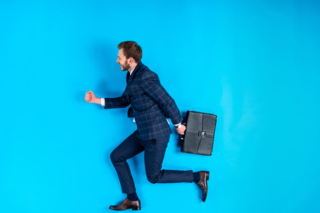 Young man in suit holding briefcase and running on blue backgroundの写真素材