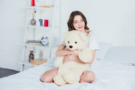 beautiful woman hugging teddy bear and sitting on bed with white beddingの写真素材