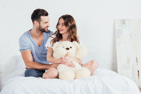 young couple hugging teddy bear while sitting on bed with white beddingの写真素材
