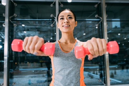 close up view of attractive asian sportswoman smiling and doing exercise with dumbbells at gymの写真素材