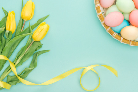 top view of yellow tulips with ribbon and easter eggs in wicker plate isolated on blue