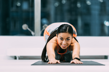 attractive asian sportswoman doing stretch exercises on fitness mat and smiling at gymの写真素材
