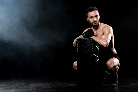 shortless man sitting and holding punching bag on black with smokeの写真素材