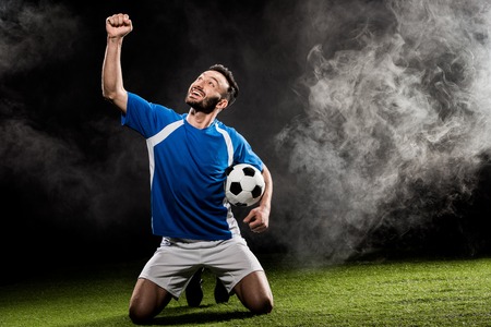 cheerful football player celebrating victory and holding ball on black with smokeの写真素材