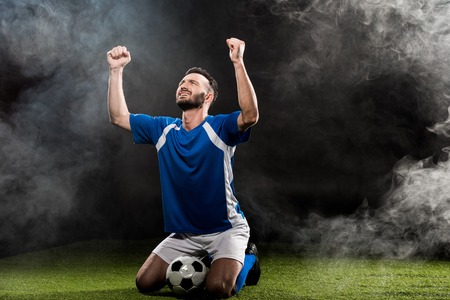 handsome football player celebrating victory while sitting on grass on black with smokeの写真素材