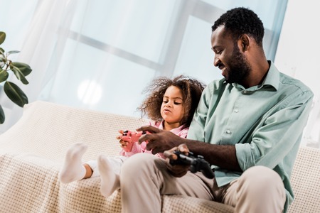 african american dad and daughter sitting on sofa and playing video game in living roomの写真素材
