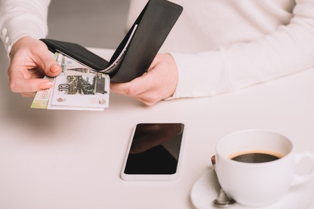 cropped shot of woman holding wallet and russian rubles banknotes above white table with cup of coffee and smartphoneの写真素材