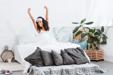 cheerful young african american woman with blindfold on forehead stretching in bed during morning time at homeの写真素材