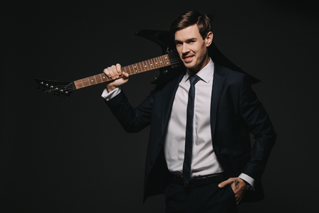 handsome businessman holding elecrtonic guitar and standing with hand in pocket isolated on blackの写真素材
