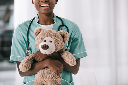 cropped view of smiling african american nurse with stethoscope holding teddy bearの写真素材