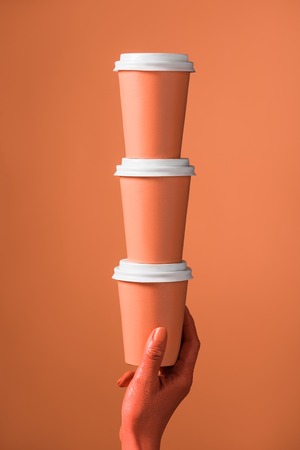 cropped view of female hand with stack of coral paper cups on coral background, color of 2019 conceptの写真素材