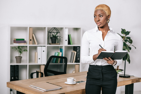pensive african american businesswoman with short hair standing near table and writing in notebookの写真素材