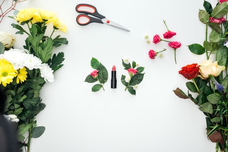 top view of pink rose buds, lipstick and scissors surrounded by flowers on white background