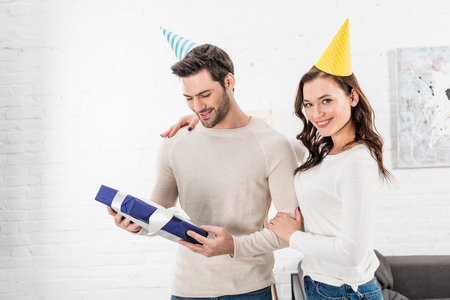 beautiful smiling couple with gift box embracing and celebrating birthday in living roomの写真素材