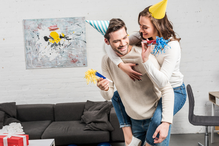 happy couple in party horns and party hats celebrating birthday while man giving piggyback ride to woman in living roomの写真素材