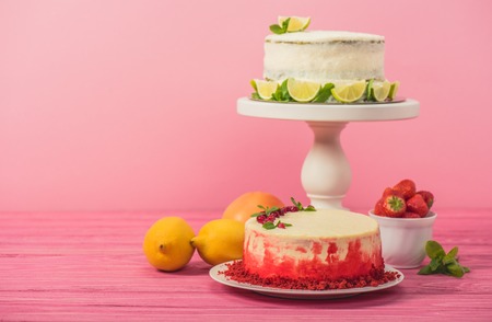cake decorated with currants and mint leaves near fruits and white cake with lemon slices on pink wooden surface isolated on pinkの写真素材