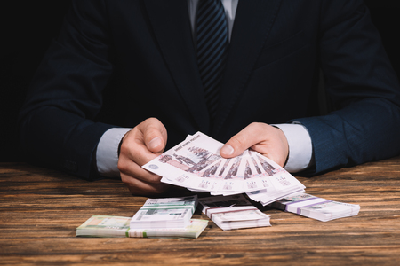 cropped shot of businessman in formal wear holding russian rubles banknotes above wooden tableの写真素材