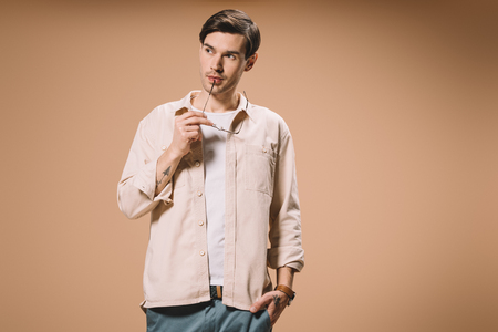 confident man holding glasses near lips and standing with hand in pocket isolated on beigeの写真素材