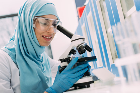 smiling female muslim scientist looking through microscope during experiment in chemical laboratoryの写真素材