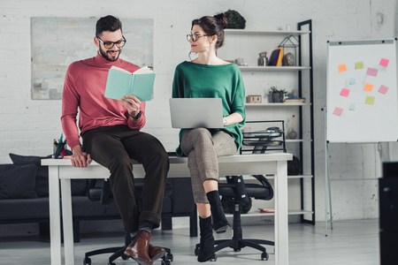 happy bearded man studying with book while sitting on table near cheerful woman with laptopの写真素材