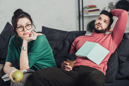 bored woman in glasses holding apple and sitting near handsome man with paper cupの写真素材