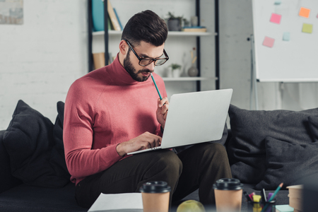 bearded man in glasses using laptop while sitting on sofaの写真素材