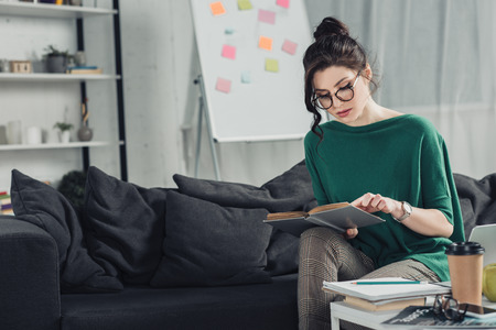 woman in glasses studying with book while sitting on sofa at homeの写真素材