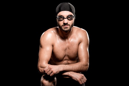 handsome swimmer sitting in swimming cap and goggles isolated on blackの写真素材