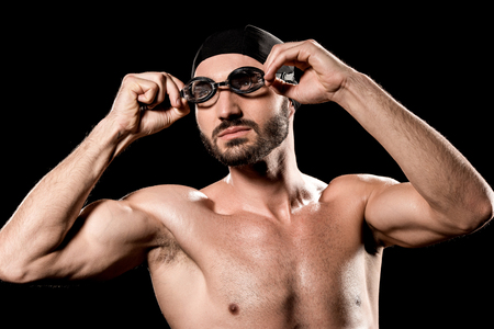 athletic sportsman standing in swimming cap and touching goggles isolated on blackの写真素材