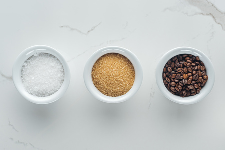 top view of bowls with salt, coffee grains and brown sugar on white surface