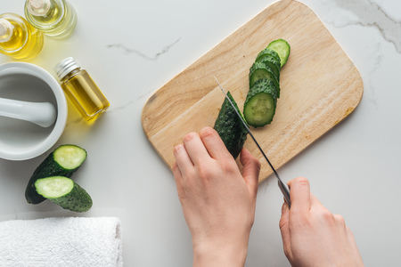 cropped view of woman cutting cucumber on wooden cutting desk, pounder and various cosmetic ingredients on white surfaceの写真素材