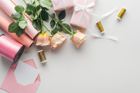 top view of roses, rolls of paper, wrapped gifts, envelope, greeting card and spools of thread on grey background