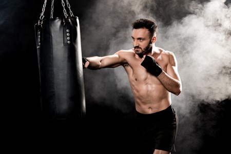 handsome shortless boxer hitting punching bag on black with smokeの写真素材