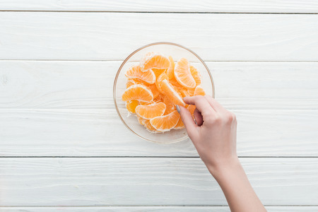 cropped view of woman taking tangerine slice from glass bowl on wooden white backgroundの写真素材