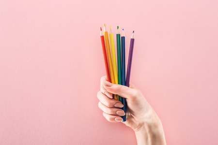 partial view of female hand with rainbow colored pencils on pink background, lgbt concept