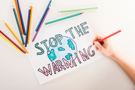 cropped view of woman writing "stop the warming" on card with globe sign, and multicolored pencils on white backgroundの写真素材