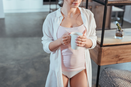 cropped image of girl in white shirt holding coffee cup during morning time at homeの写真素材