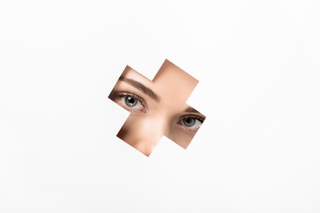partial view of attractive girl looking at camera through cross shaped hole on whiteの写真素材