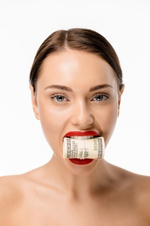 beautiful young woman holding rolled dollar banknotes in teeth and looking at camera isolated on...