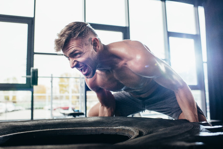 handsome shirtless sportsman lifting tire and screaming in gym