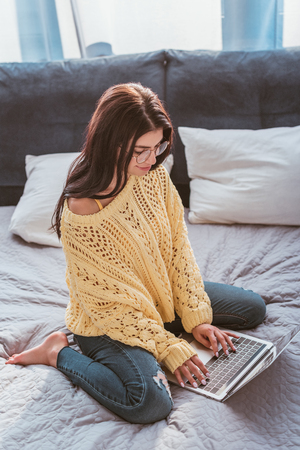 young female freelancer in eyeglasses working on laptop while sitting on bed at homeの写真素材