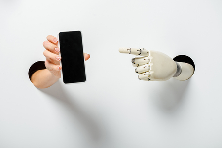 cropped image of woman holding smartphone with blank screen and robot pointing on it through holes on whiteの写真素材